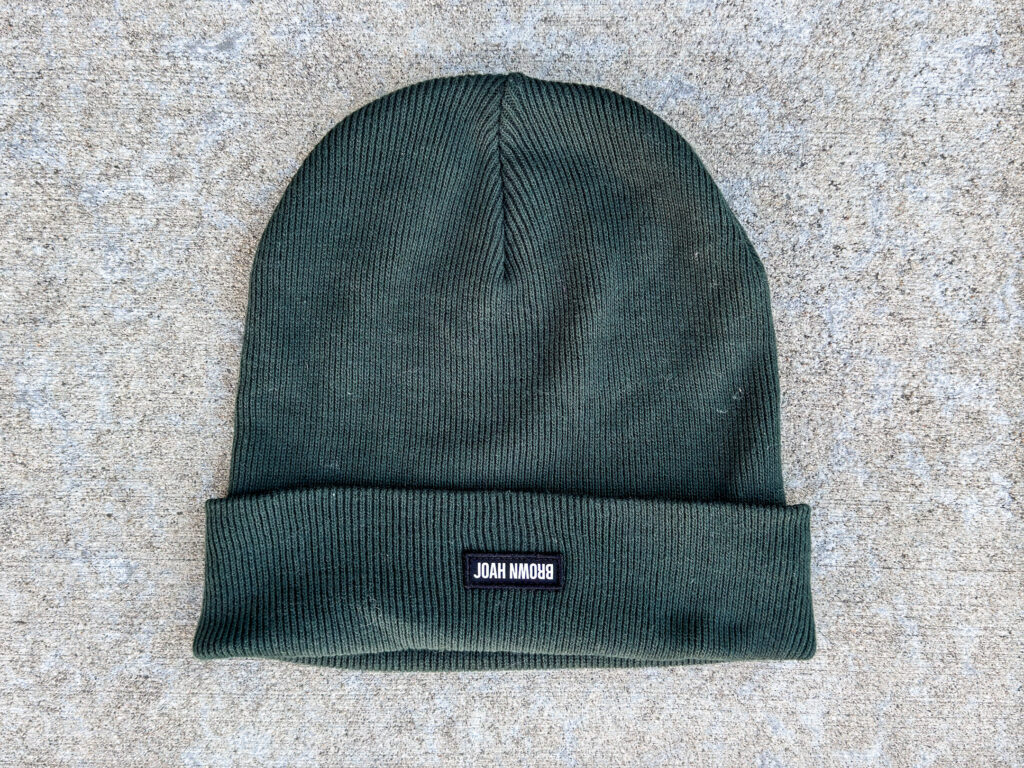 A dark green knit beanie with a folded brim, featuring a small black patch with white text that reads "JOAH BROWN."