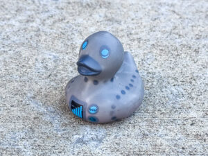 A small rubber duck with a metallic gray body, blue glowing-like eyes, and futuristic circuit-like markings, resembling a robotic or cybernetic duck.