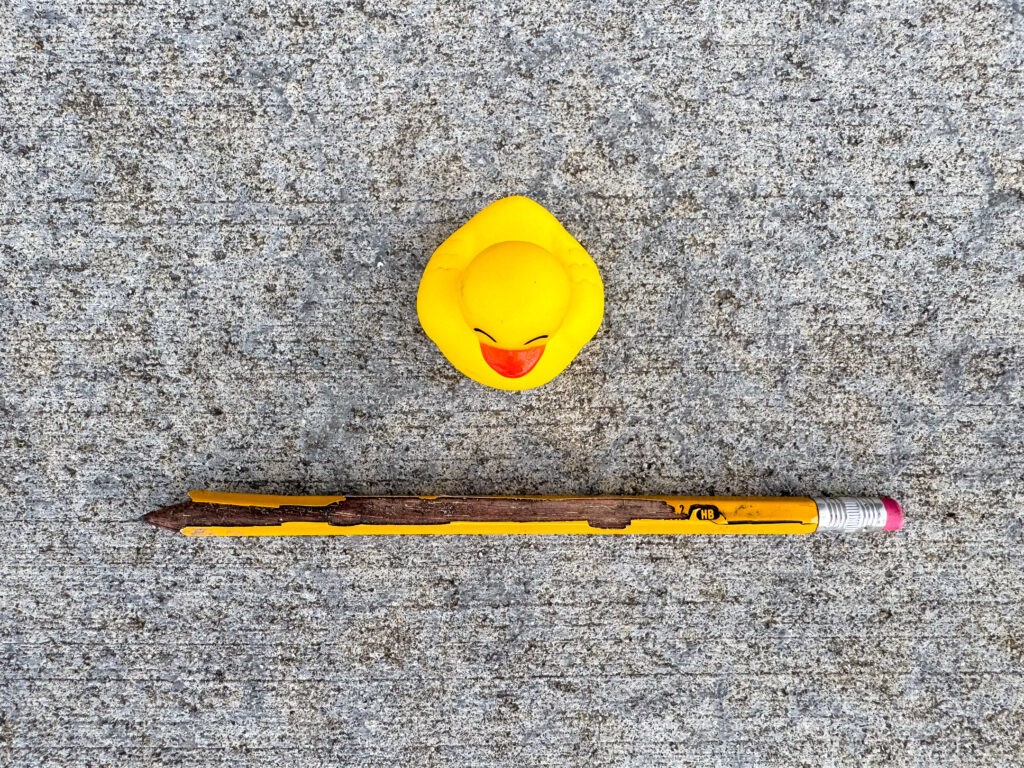 A small yellow rubber duck with an orange beak and a heavily worn HB pencil with chipped yellow paint and pink eraser.