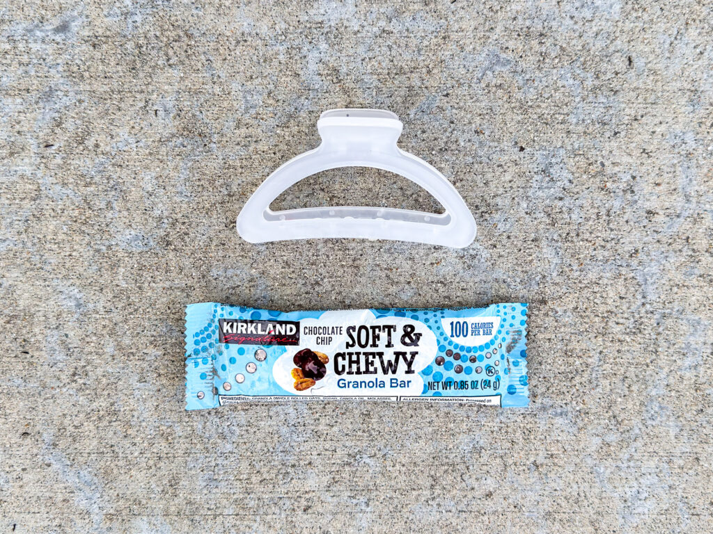 A translucent plastic hair clip positioned above a Kirkland Signature Soft & Chewy chocolate chip granola bar in its packaging.