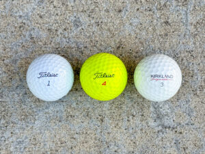Three golf balls in a row: white Titleist 1, yellow Titleist 4, and white Kirkland Signature 3.