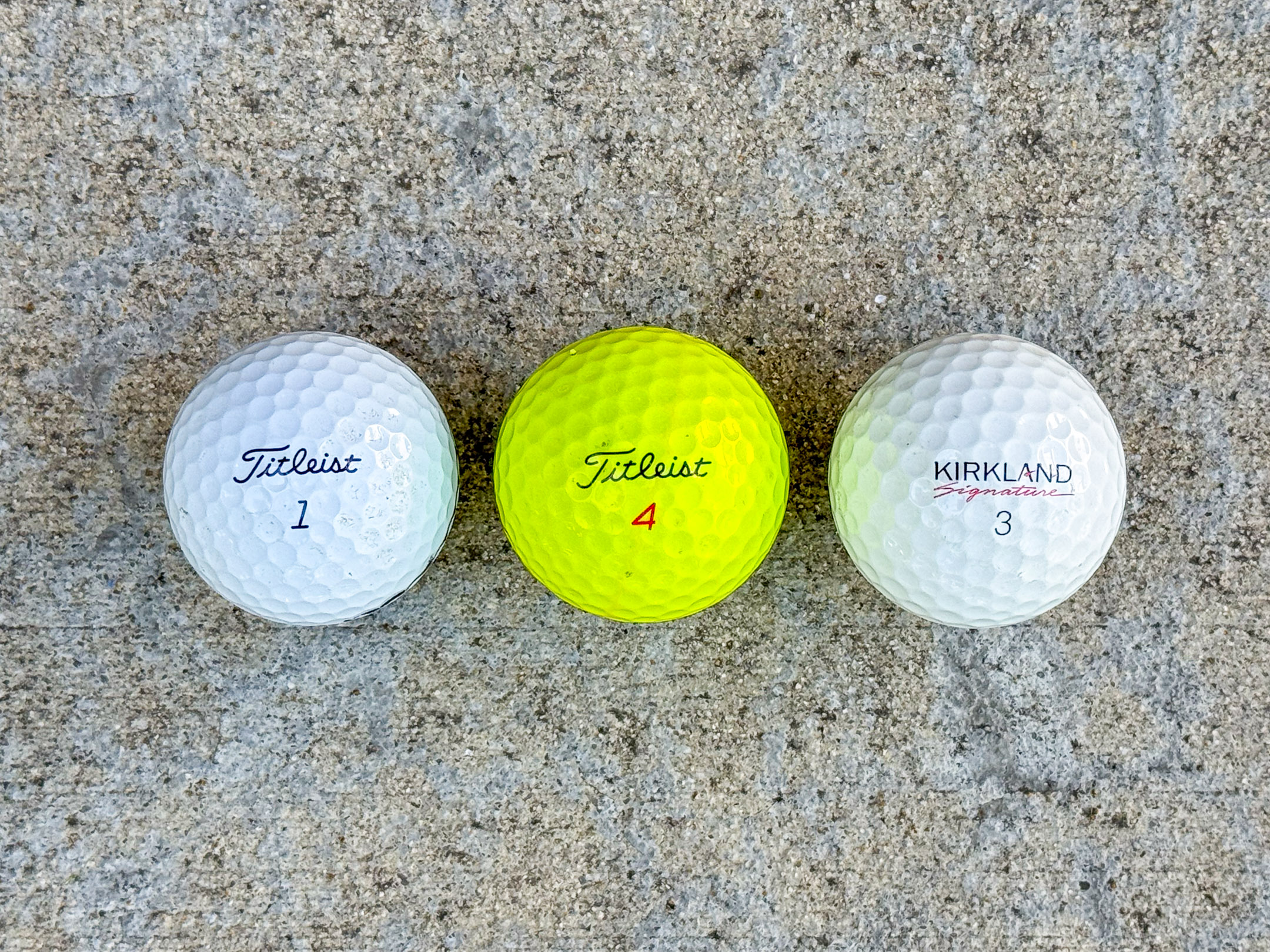 Three golf balls in a row: white Titleist 1, yellow Titleist 4, and white Kirkland Signature 3.