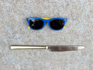 Small blue plastic sunglasses with yellow arms placed above a stainless steel serrated table knife marked “STAINLESS CHINA J".