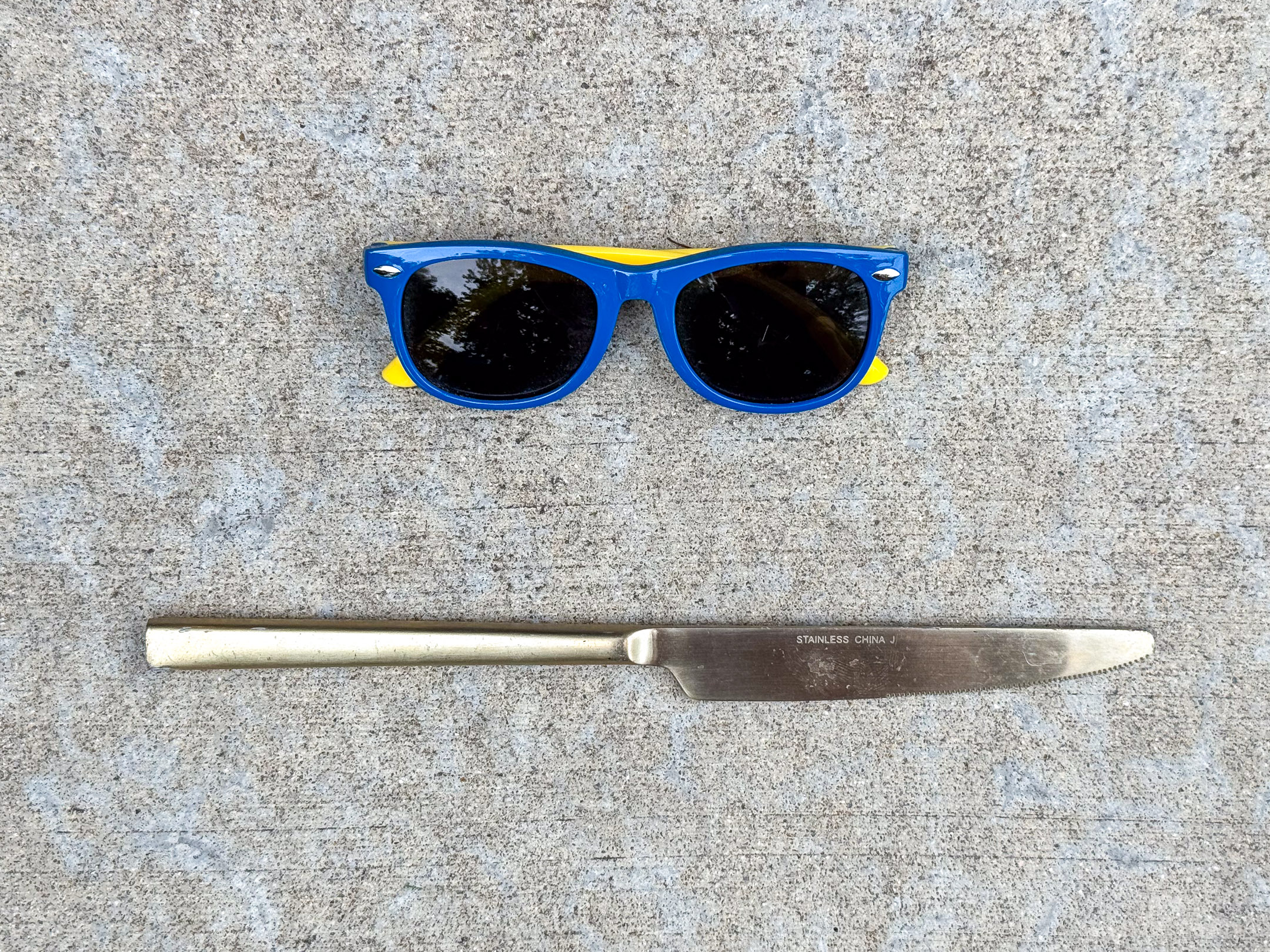 Small blue plastic sunglasses with yellow arms placed above a stainless steel serrated table knife marked “STAINLESS CHINA J".