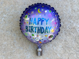 A round, metallic purple birthday balloon with a space theme and large “HAPPY BIRTHDAY” text, anchored by a large, pitted lug nut.