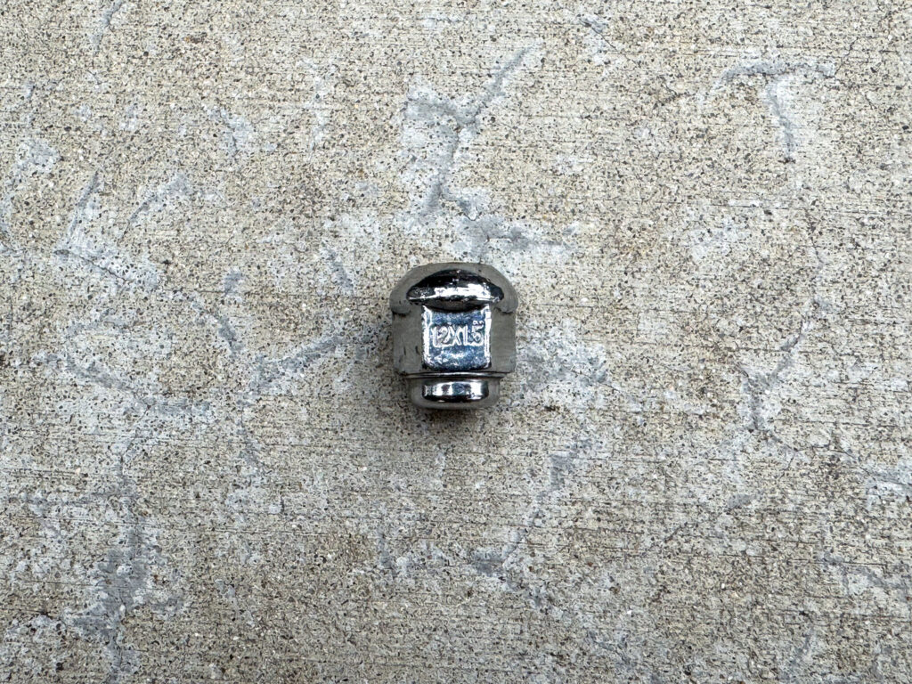 A shiny chrome acorn lug nut with “12x1.5” engraved on one of the flats.