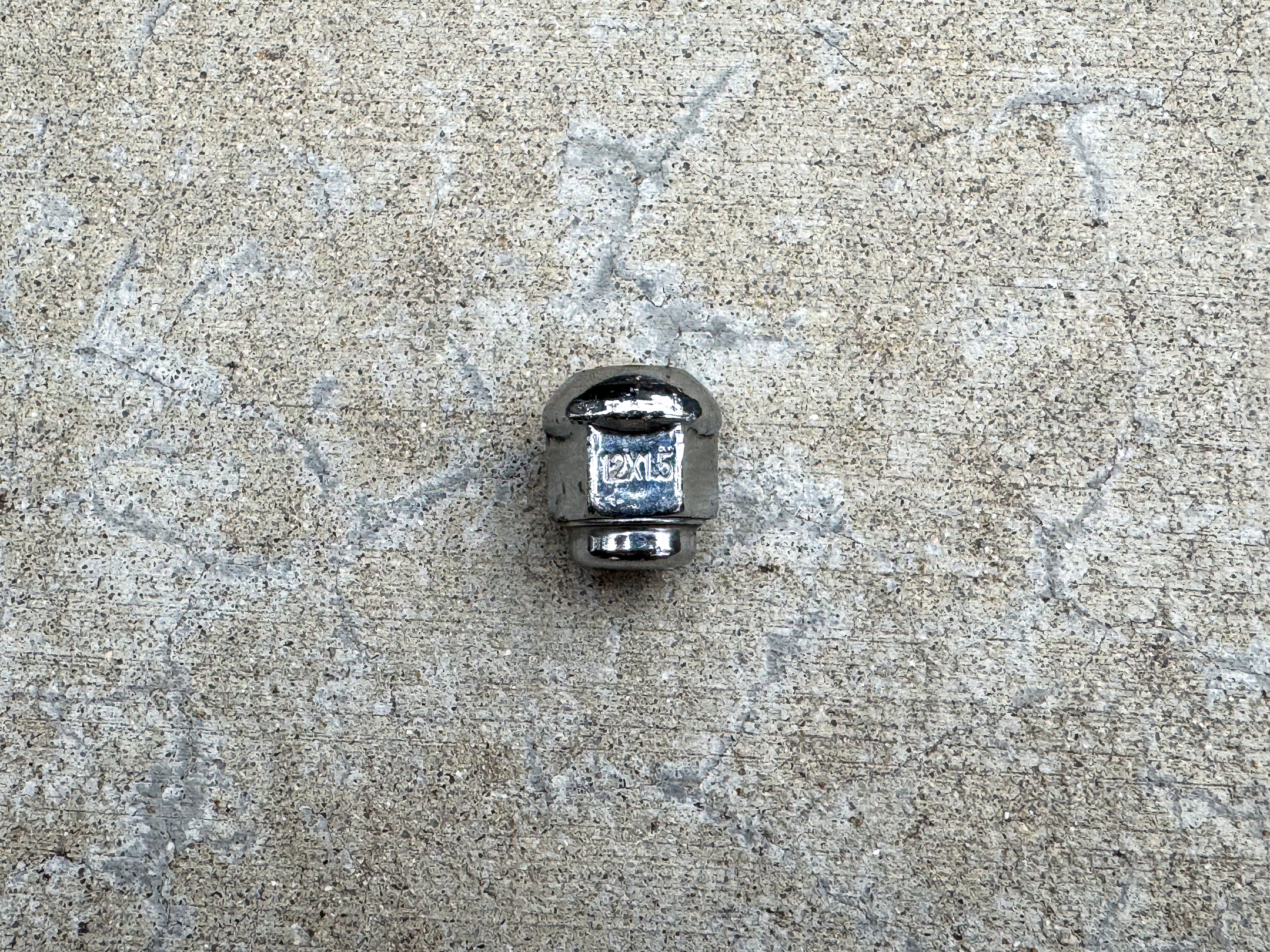 A shiny chrome acorn lug nut with “12x1.5” engraved on one of the flats.