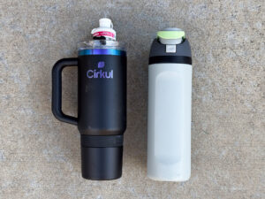 A matte black Cirkul bottle with a handle with a white “Mango Passion Fruit” flavor cartridge next to a tall light gray bottle with a dark gray lid and light green flip-top button.