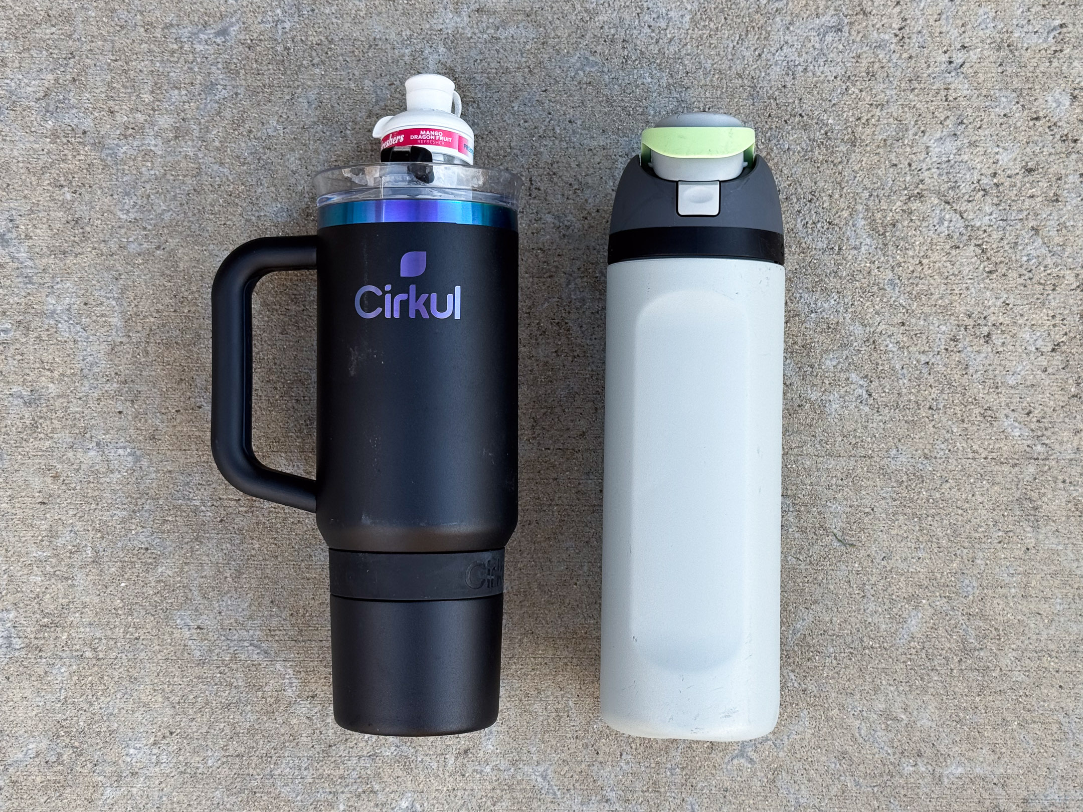 A matte black Cirkul bottle with a handle with a white “Mango Passion Fruit” flavor cartridge next to a tall light gray bottle with a dark gray lid and light green flip-top button.