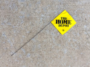 Yellow survey flag on a thin metal wire with black stencil text reading THE HOME DEPOT, lying diagonally.