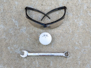 Black safety glasses above a white Snake Eyes 2 golf ball and a chrome 12 mm Craftsman combination wrench, arranged vertically.