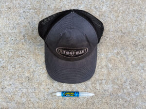 Black Stoneway trucker hat with a mesh back and stitched front patch, placed above a blue Juicy Drop candy pen.