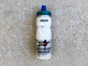Scuffed white CamelBak cycling water bottle with a teal cap, blue nozzle, subtle hexagon pattern, and Bike’s Edge, Kaysville, UT logo.