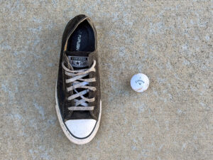 Worn black Converse All Star sneaker with gray laces and a dirty white rubber toe beside a scuffed white Callaway 3 golf ball.