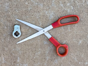 Red-handled scissors with open steel blades beside a scuffed chrome Harley-Davidson side stand stop marked “DOWN SIDE”, with a square center hole.