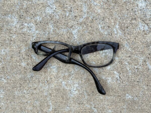 Broken black rectangular reading glasses with one loose or missing lens, twisted temples, scratched dark frames, and “+2.50” marking visible on one arm.