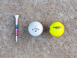 White golf tee with an American flag pattern beside a white Callaway 2 golf ball and a bright yellow Pinnacle Practice golf ball, both lightly scuffed.