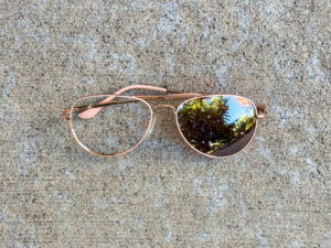 Rose-gold aviator glasses with pink temple tips, one clear lens, and one dark mirrored lens reflecting trees and sky.
