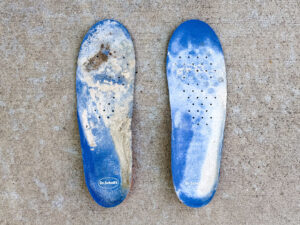 Two blue Dr. Scholl’s size 13 insoles, heavily worn and coated with dried gray-white construction residue.