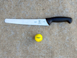 Long Mercer serrated bread knife with a black textured handle beside a bright yellow Pinnacle Practice golf ball.